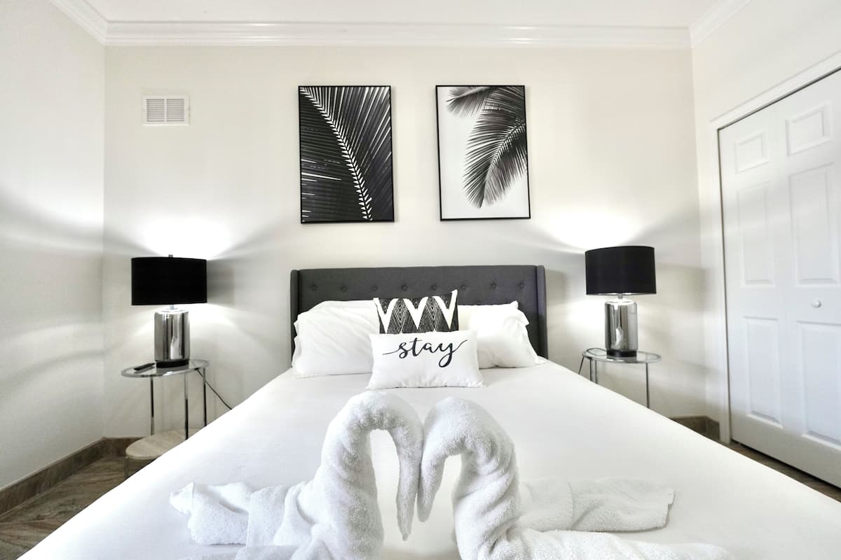 A neatly made bed with white linens, two wall-mounted picture frames, two bedside lamps, and a pillow with the word "stay".