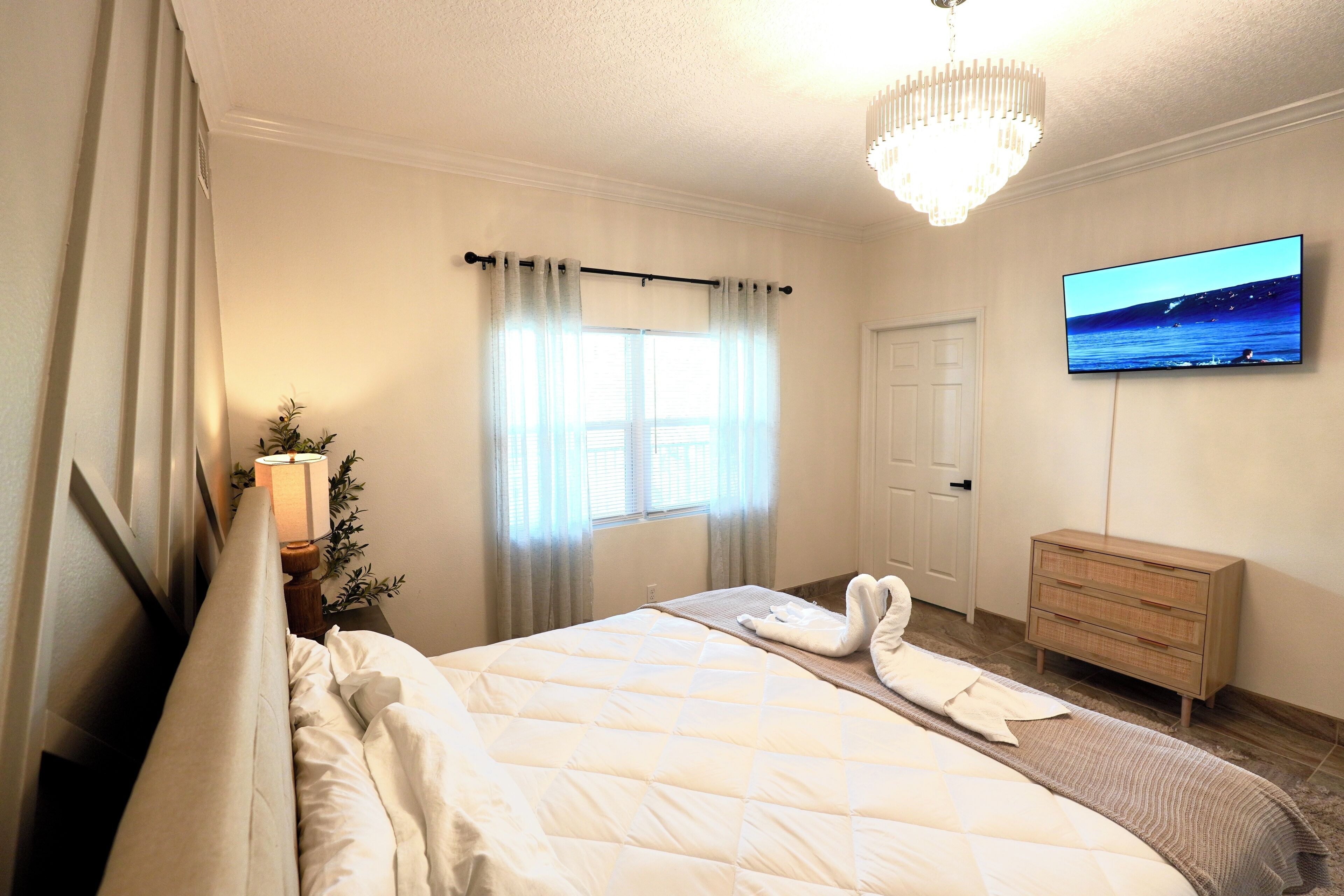 A hotel room with a bed, a TV on the wall, a dresser, and a window with curtains.