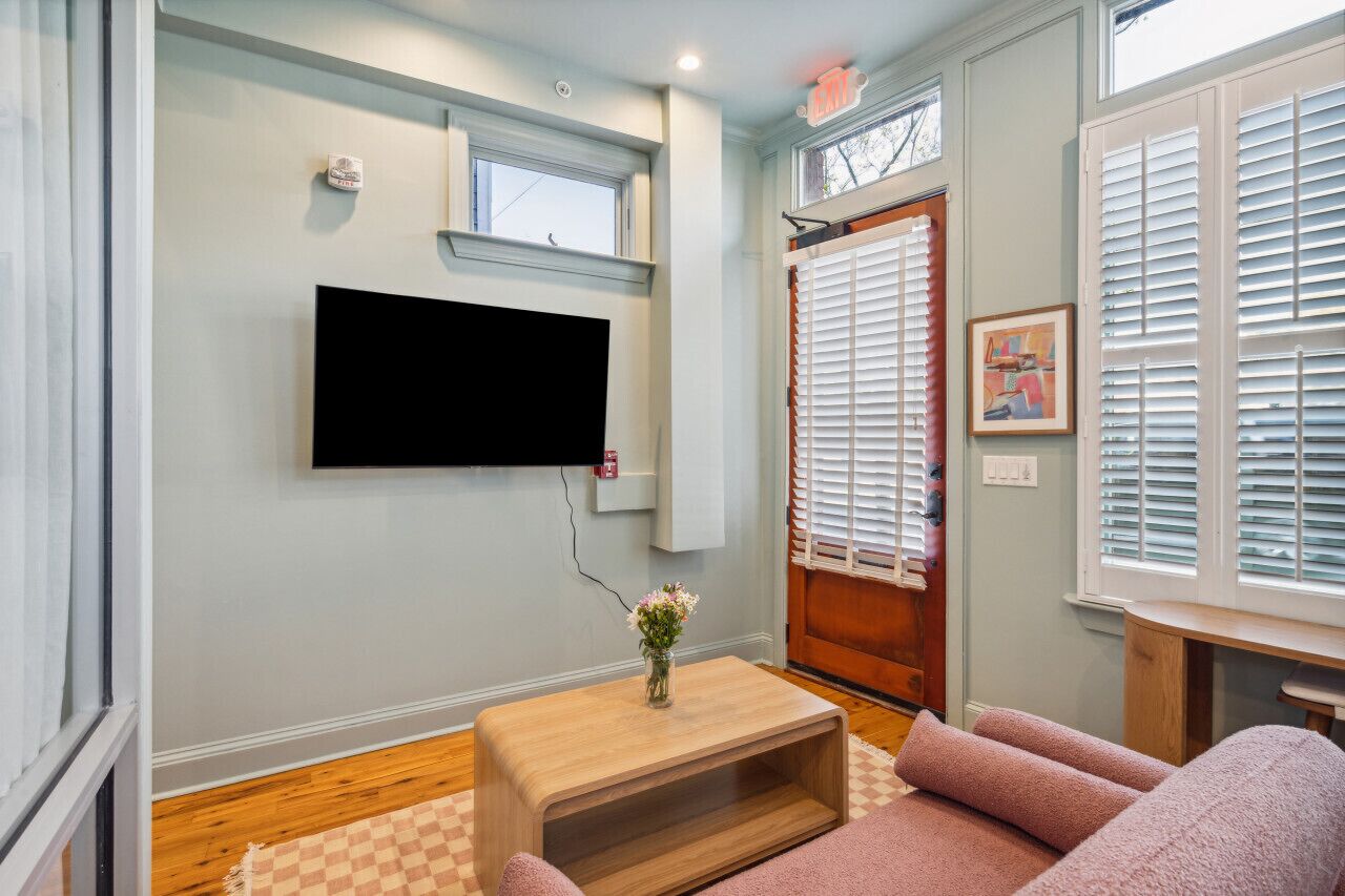 Una sala de estar con un televisor de pantalla plana montado en la pared, una mesa de centro de madera con un jarrón de flores y un sofá rosa.