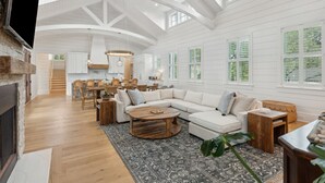 Ein geräumiges Wohnzimmer mit einer großen, L-förmigen Couchgarnitur, einem Holztisch und einem Kamin. Es gibt einen Essbereich mit einem Holztisch und Stühlen, und im Hintergrund ist eine Küche zu sehen.