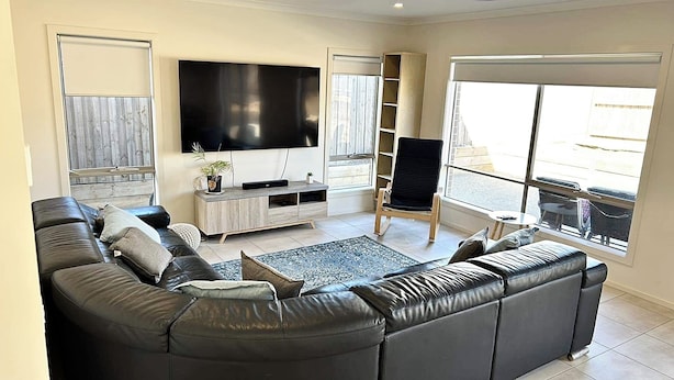 A modern living room with a large black leather sofa, a flat-screen TV mounted on the wall, a wooden entertainment unit, and a view through large windows. - Spacious Coastal Oasis - 10 min to Torquay, 10 min to Geelong ! (Mount Duneed)