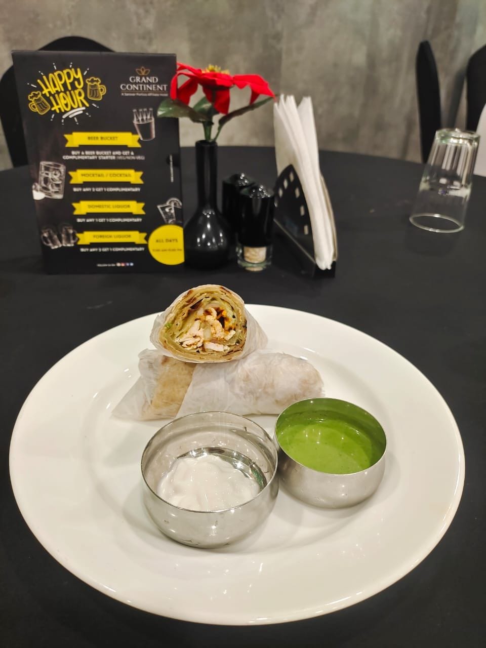 A plate with a wrap, a small bowl of white sauce, and a green sauce on a white plate. A black box with yellow text and a red flower is in the background.