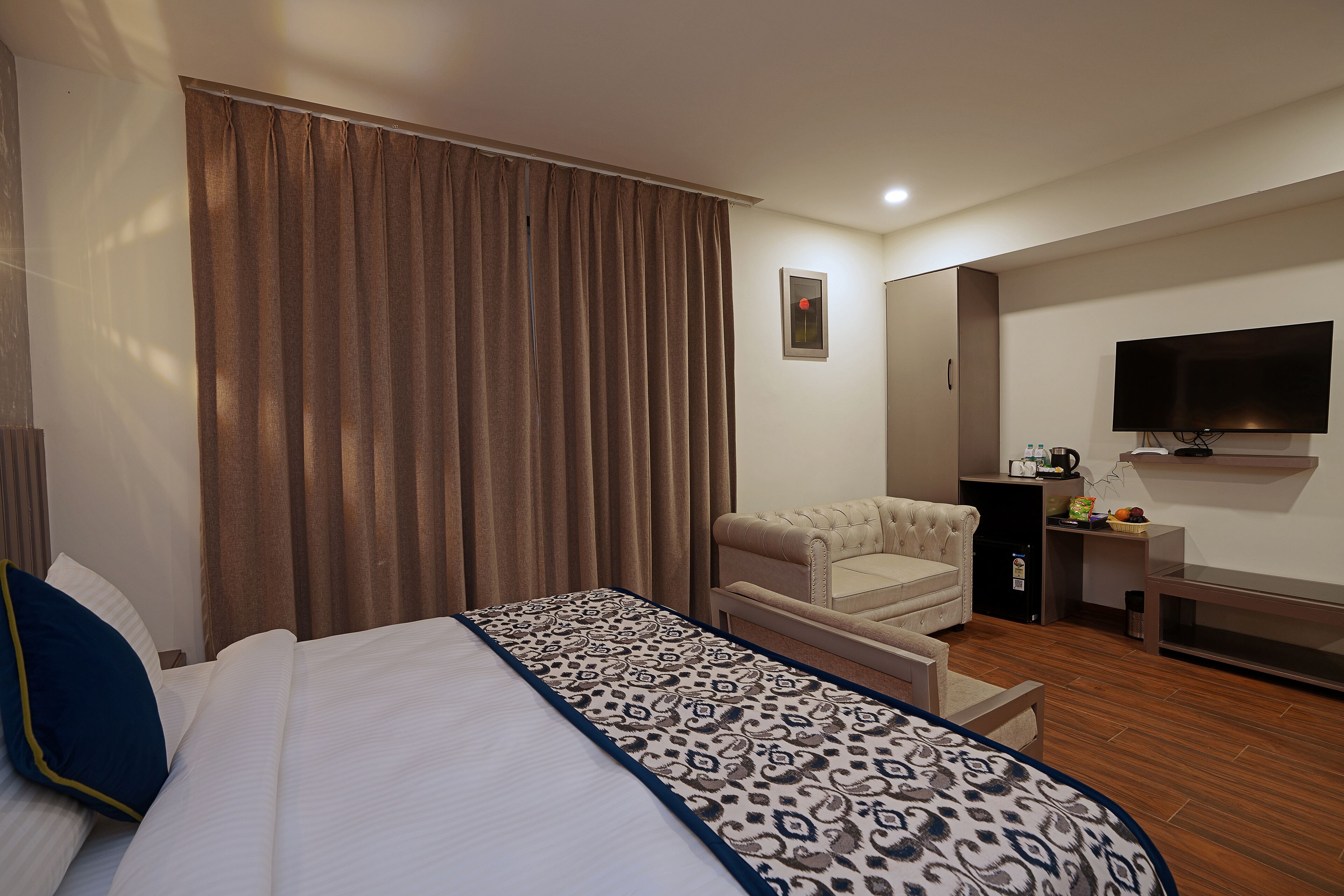 A hotel room with a bed, a sofa, a TV, and a desk with a kettle and fruit.