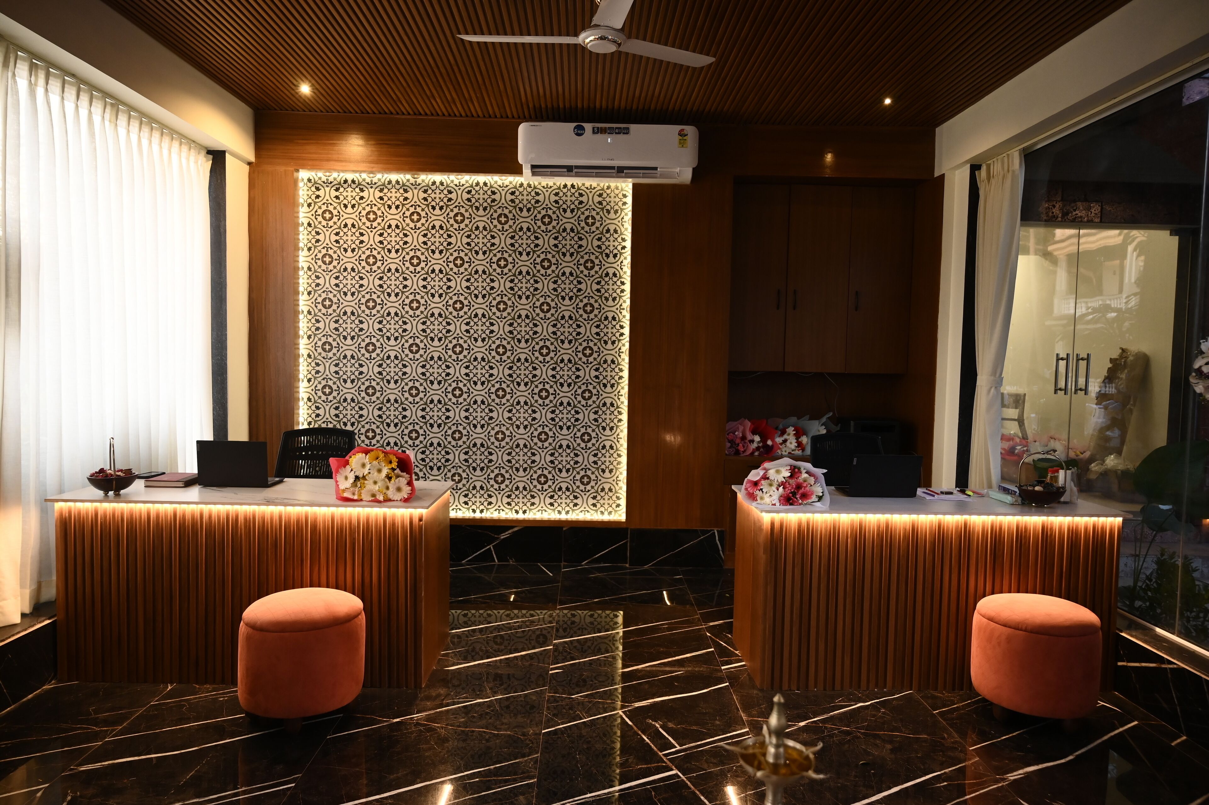 A reception area with a modern design, featuring a patterned wall, a ceiling fan, and a reception desk with a laptop and decorative items.