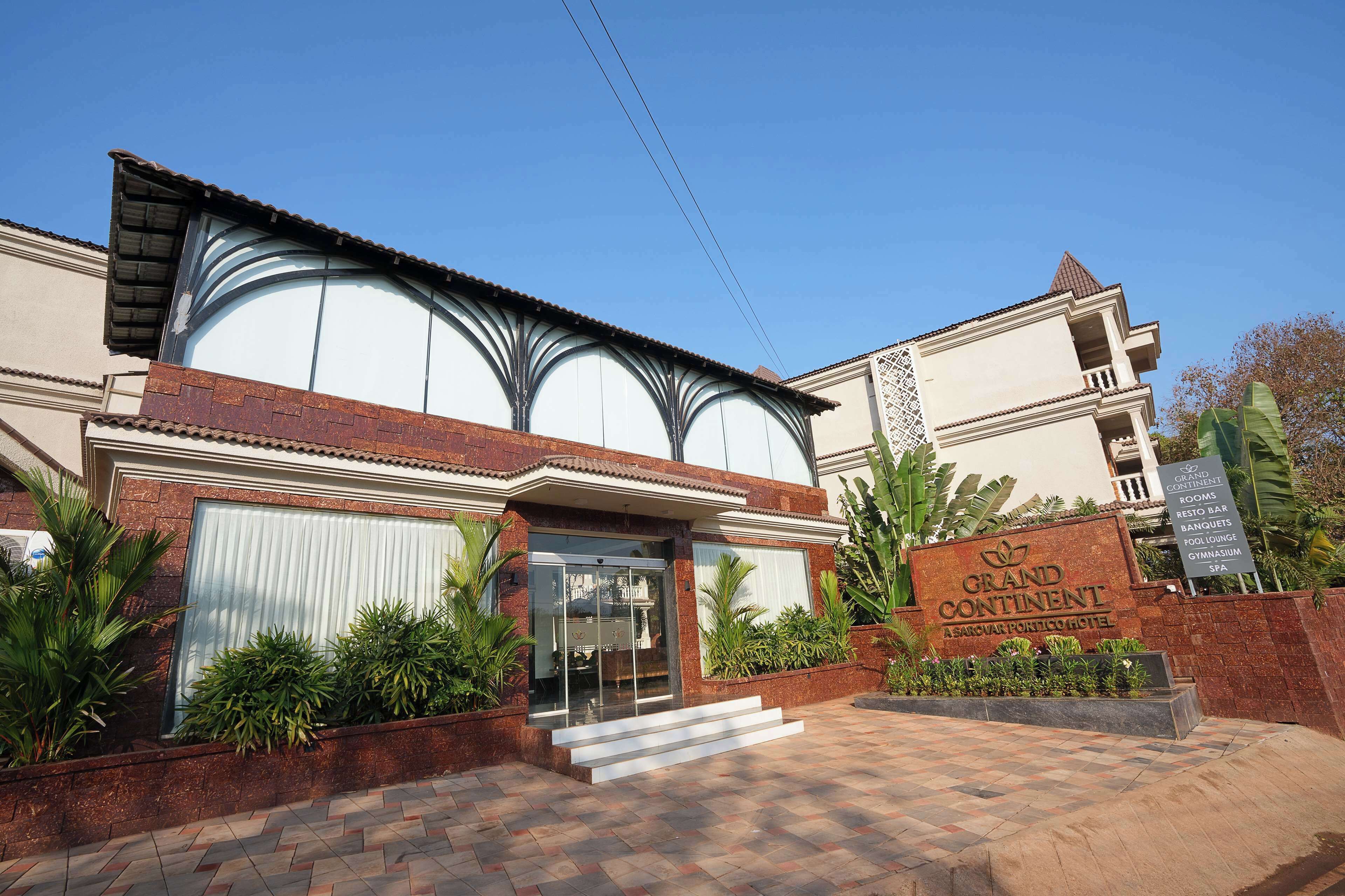 A hotel entrance with a sign reading "Grand Continental A Sarovar Portico Hotel," a gated entrance, and a paved driveway.