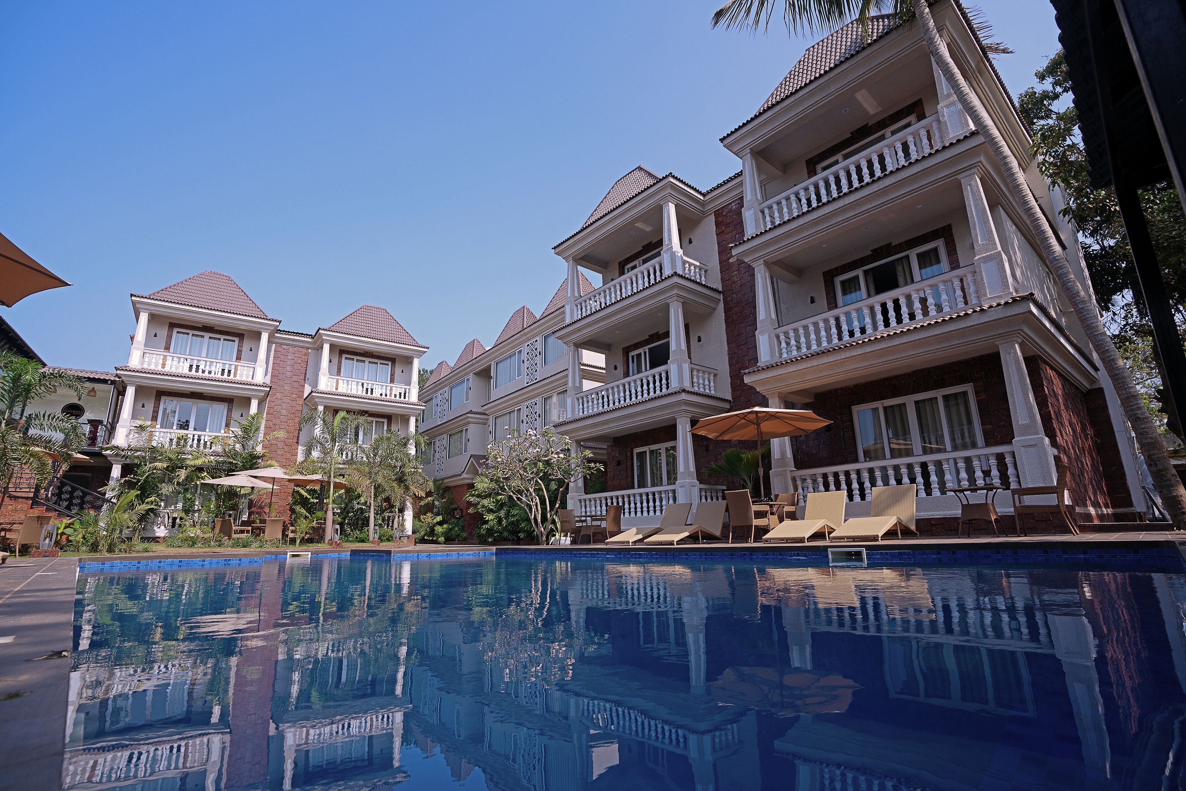 Un hotel de varios pisos con piscina, mobiliario exterior y un cielo azul despejado.