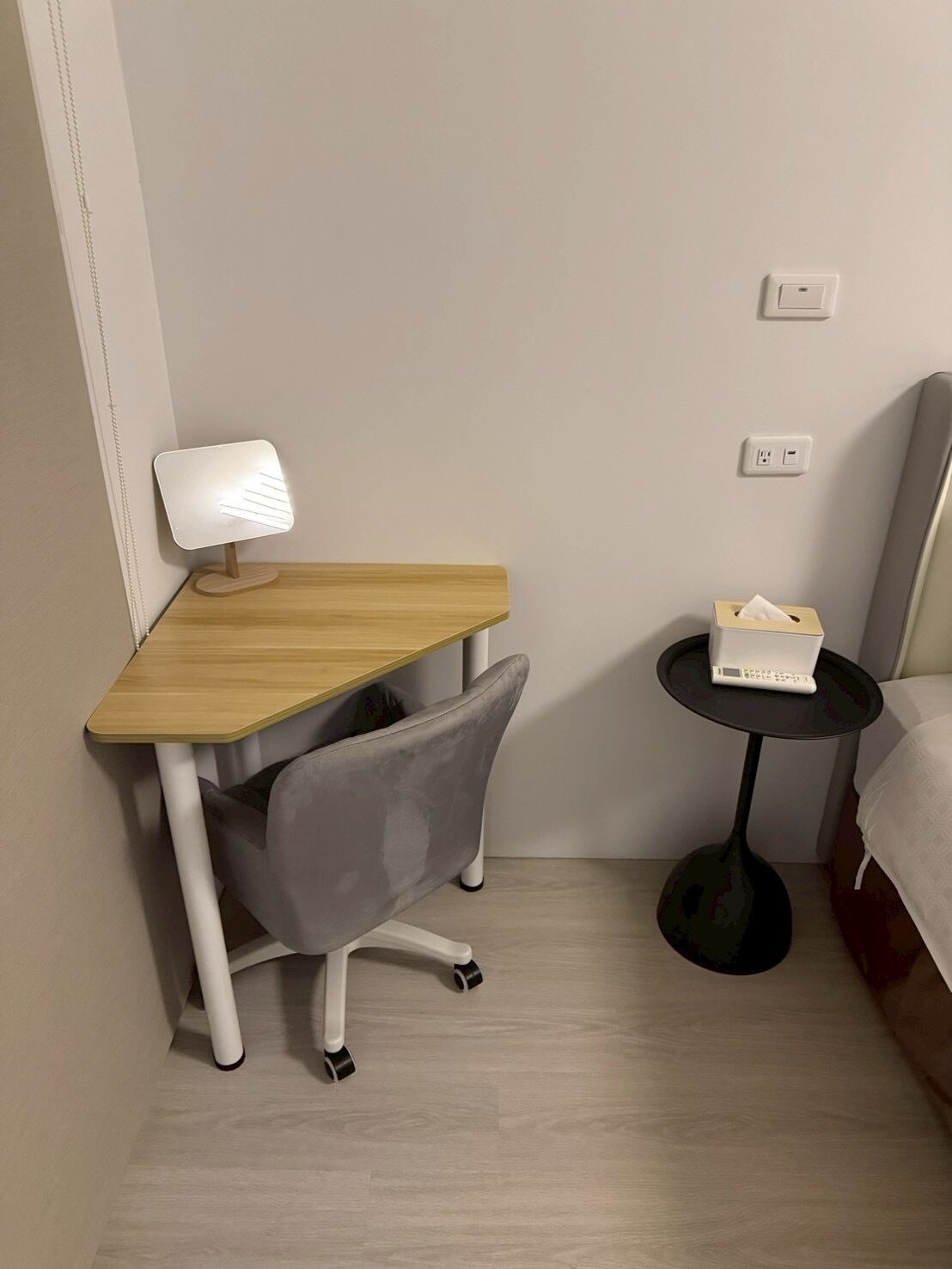 A small, modern room with a wooden desk, a grey office chair, a bedside table with a lamp, and a bed.