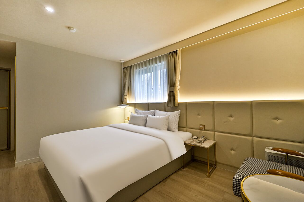 A hotel room with a large bed, a bedside table with a lamp, a headboard, and a small seating area with a chair and a table.