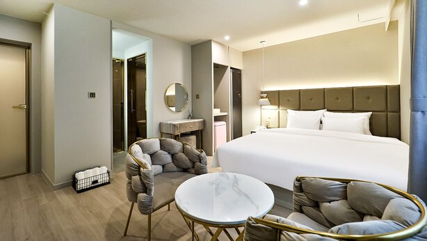A hotel room with a large bed, a round marble table, two armchairs, a small round table, a mirror, and a bathroom visible through an open door. - Den Basta Hotel Daeyeon (Busan)