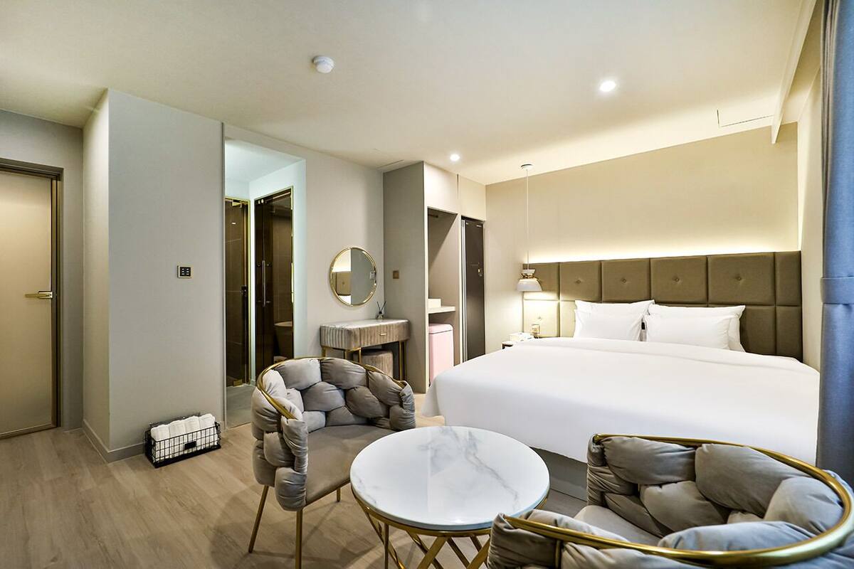 Una habitación de hotel con una cama grande, una mesa redonda de mármol, dos sillones, una pequeña mesa redonda, un espejo y un baño visible a través de una puerta abierta.