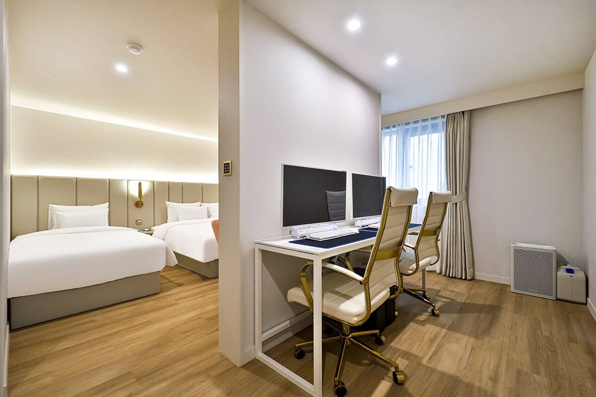 Una habitación de hotel con dos camas, un escritorio con dos monitores y una ventana con cortinas.