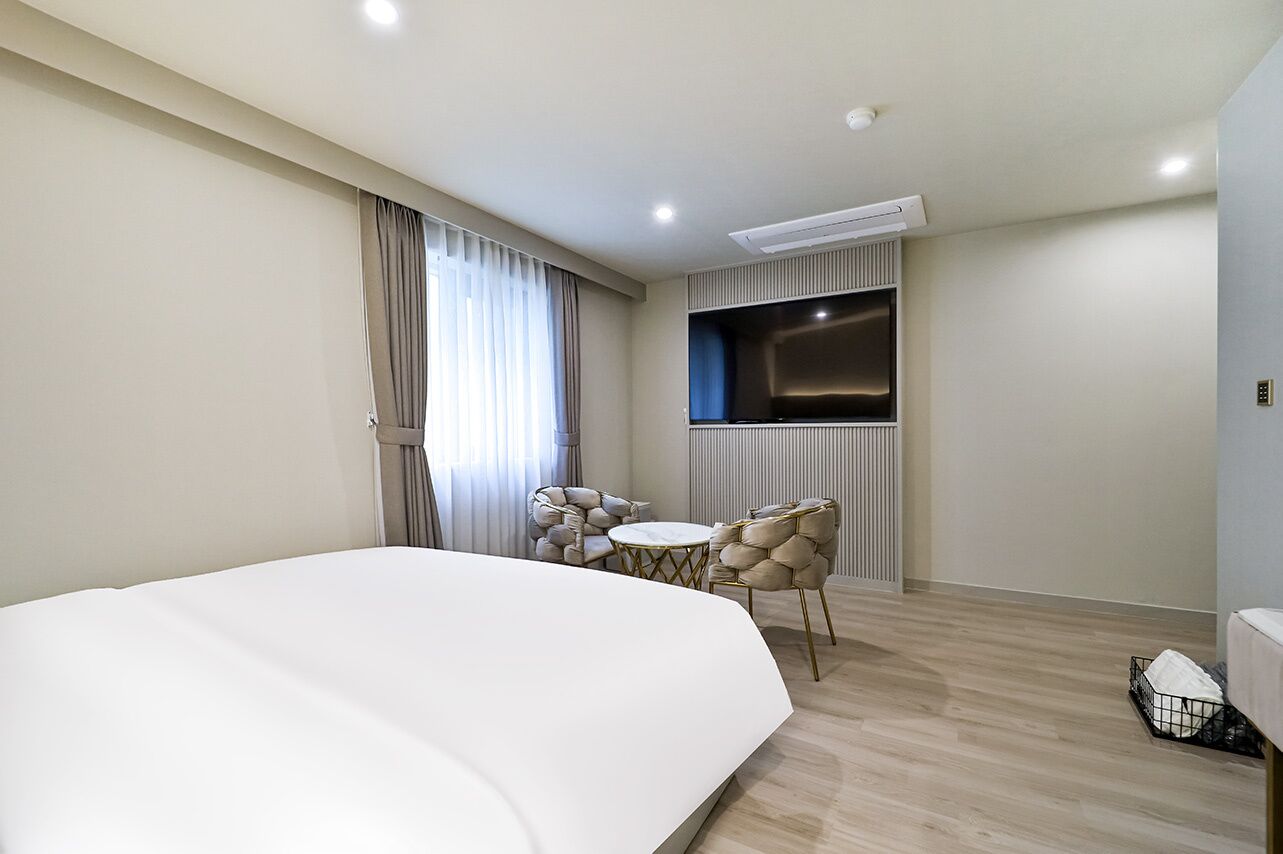 A hotel room with a large bed, a TV mounted on the wall, a small round table with chairs, and a window with curtains.