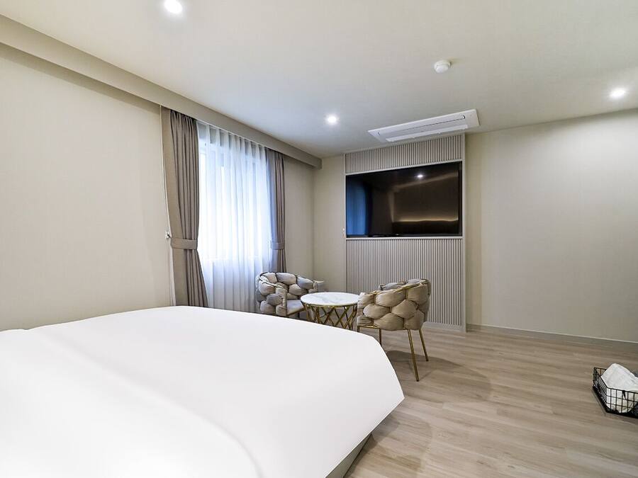 A hotel room with a large bed, a TV mounted on the wall, a small round table with chairs, and a window with curtains.