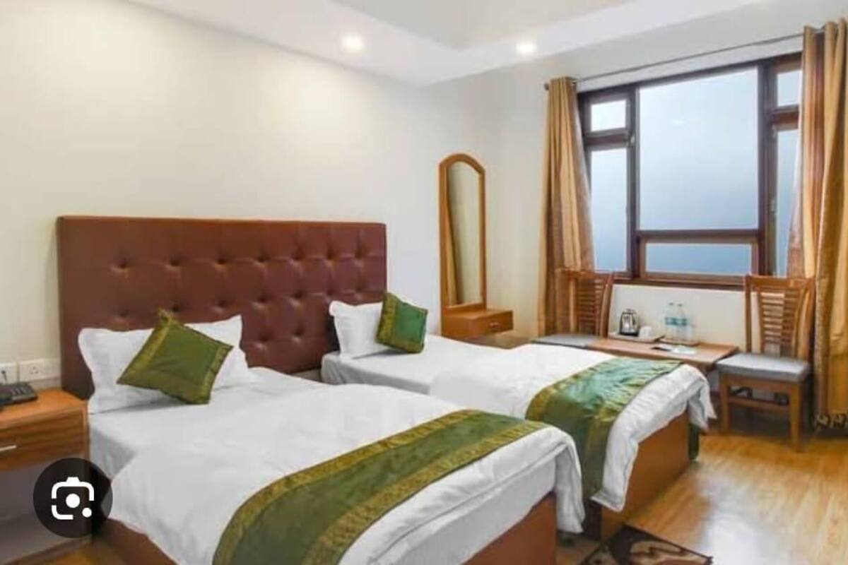 A hotel room with two beds, a wooden headboard, a mirror, a window with curtains, and a small table with a chair.