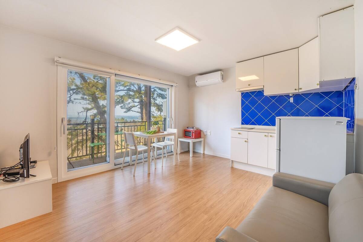 A modern kitchen with a blue backsplash, a dining area with a table and chairs, and a large sliding glass door leading to a balcony with a view of trees and the sea.