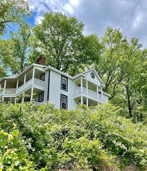 A two-story house with a balcony on the second floor, surrounded by lush greenery.