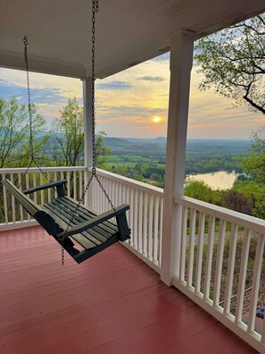 A porch with a swing chair, white railings, and a view of a landscape with trees and a body of water.