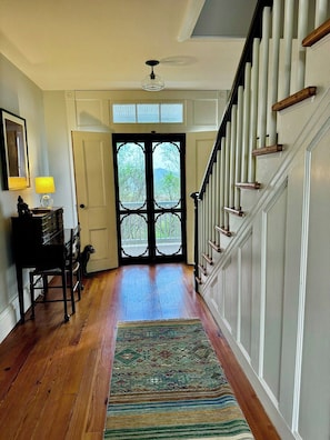 A hallway with a wooden floor, a patterned rug, a desk with a lamp, and a staircase with white railings.