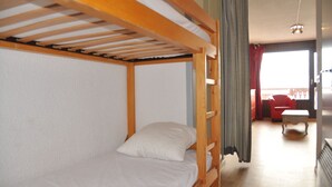 Un lit superposé avec une structure en bois et du linge de lit blanc, une petite table avec une chaise blanche, et une vue sur un balcon avec des rideaux rouges.