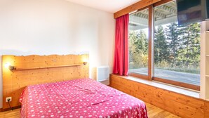Um quarto com cabeceira de madeira, uma cama com colcha estampada de flores vermelhas, uma janela com cortina vermelha e uma TV fixada na parede.