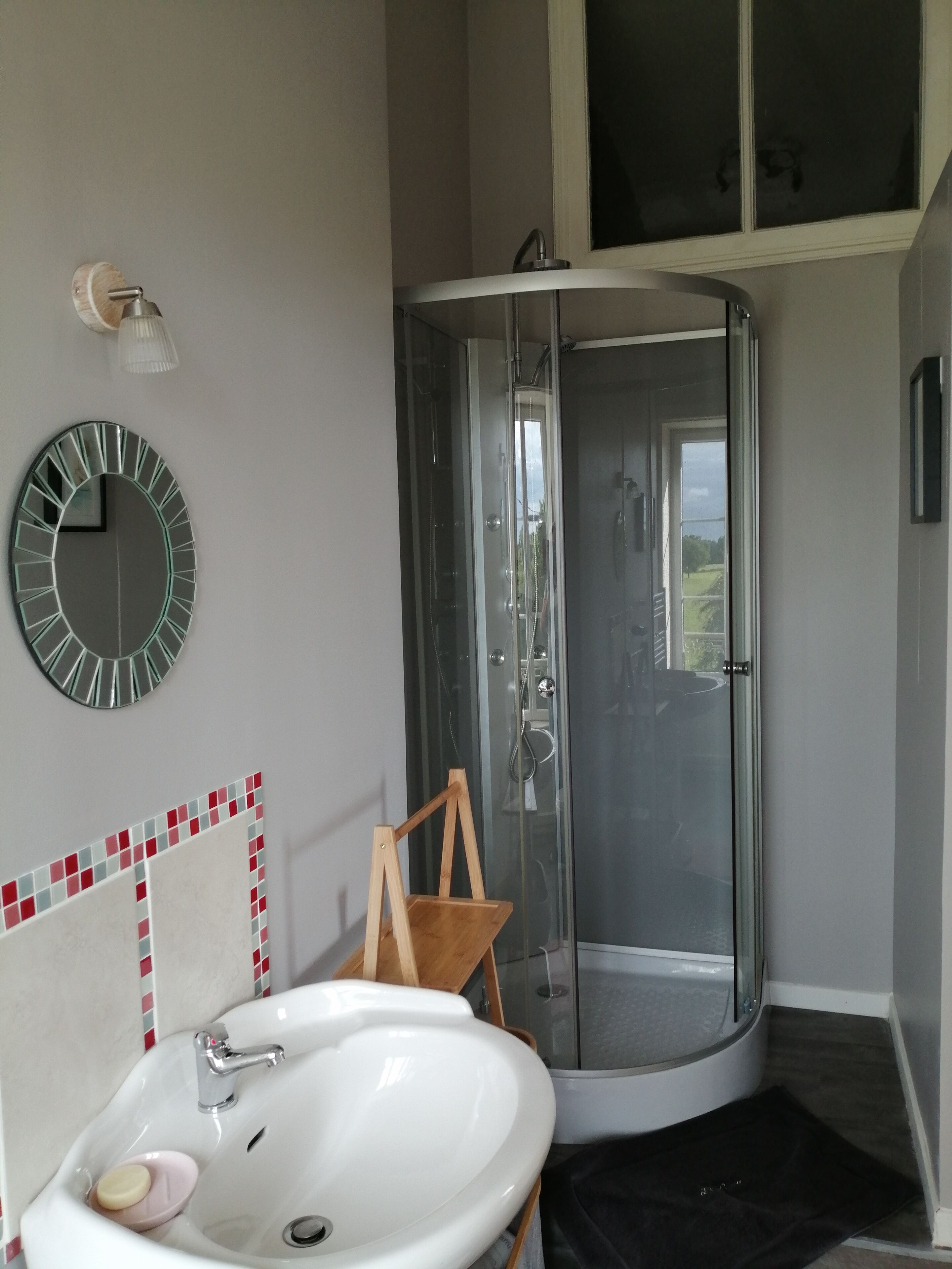 Une salle de bain avec une douche à parois de verre, un lavabo blanc avec un porte-savon, une chaise en bois et un miroir au mur.