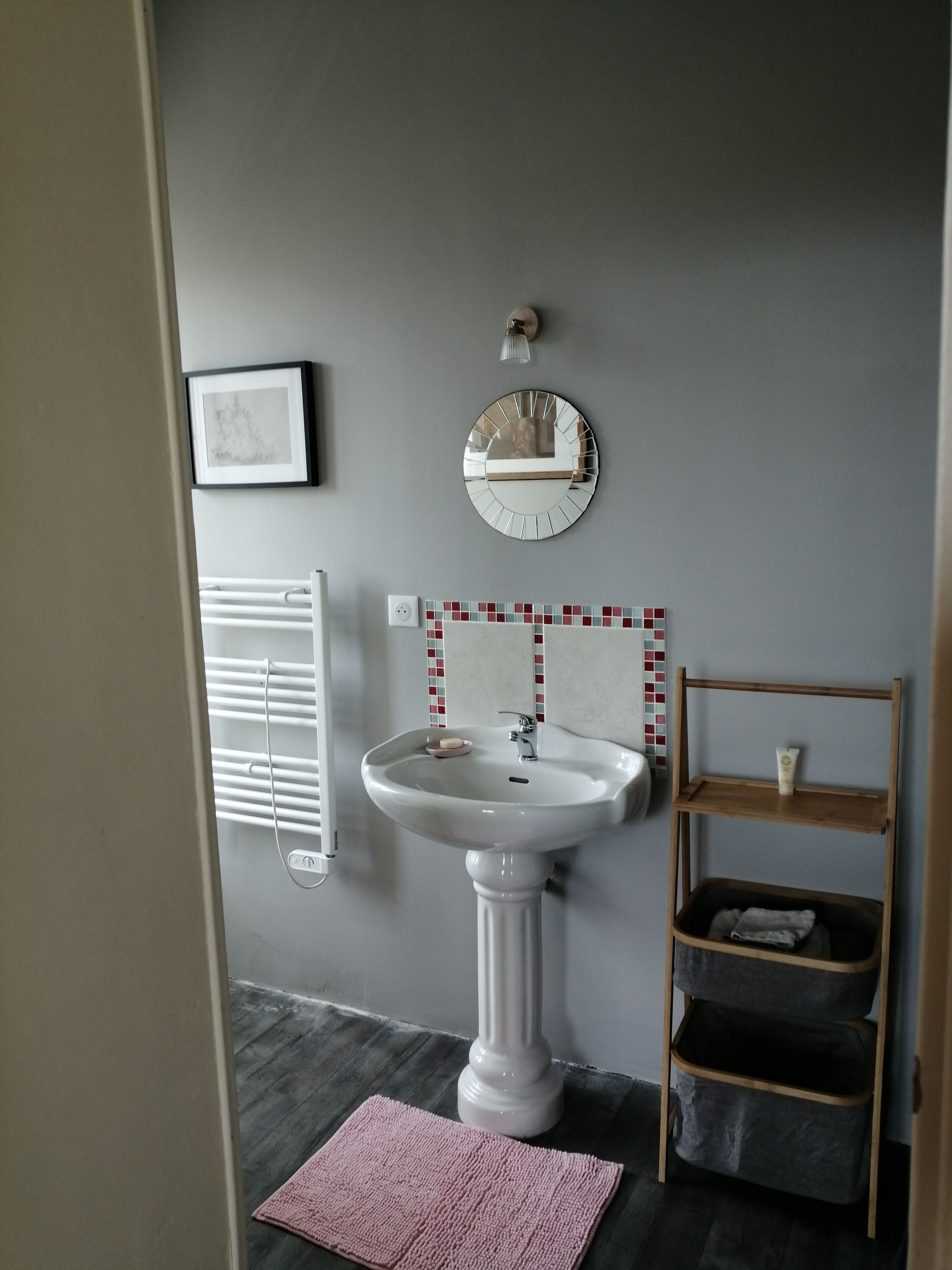Ein Badezimmer mit einem Standwaschbecken, einem runden Spiegel, einer kleinen Ablage mit Pflegeprodukten, einem Handtuchhalter und einem gerahmten Bild an der Wand.