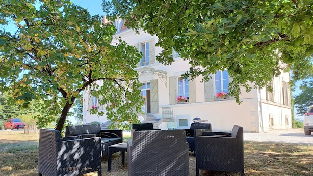 An outdoor seating area with wicker furniture, a white building with shutters in the background, and a tree providing shade.