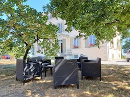 An outdoor seating area with wicker furniture, a white building with shutters in the background, and a tree providing shade.