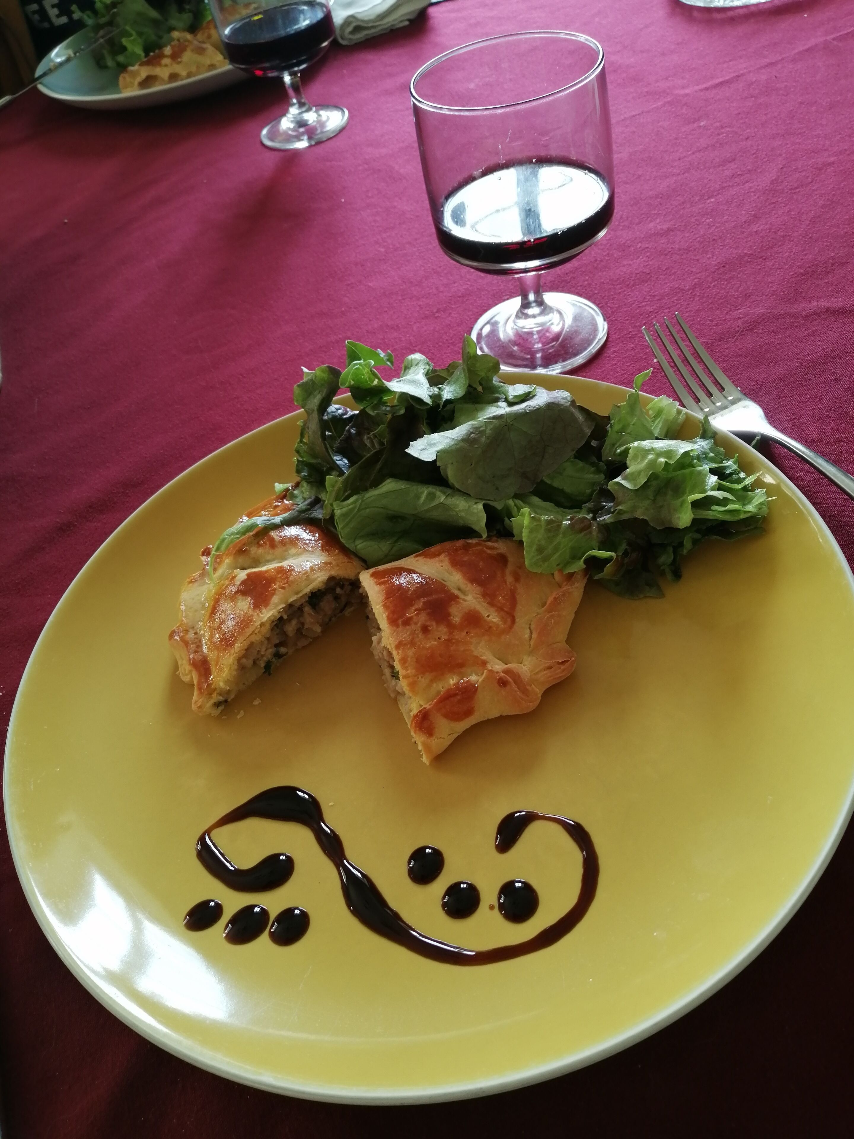 Une assiette jaune sur laquelle sont disposés une pâtisserie et une salade, un verre de vin rouge et une fourchette, le tout sur une table recouverte d’une nappe rouge.