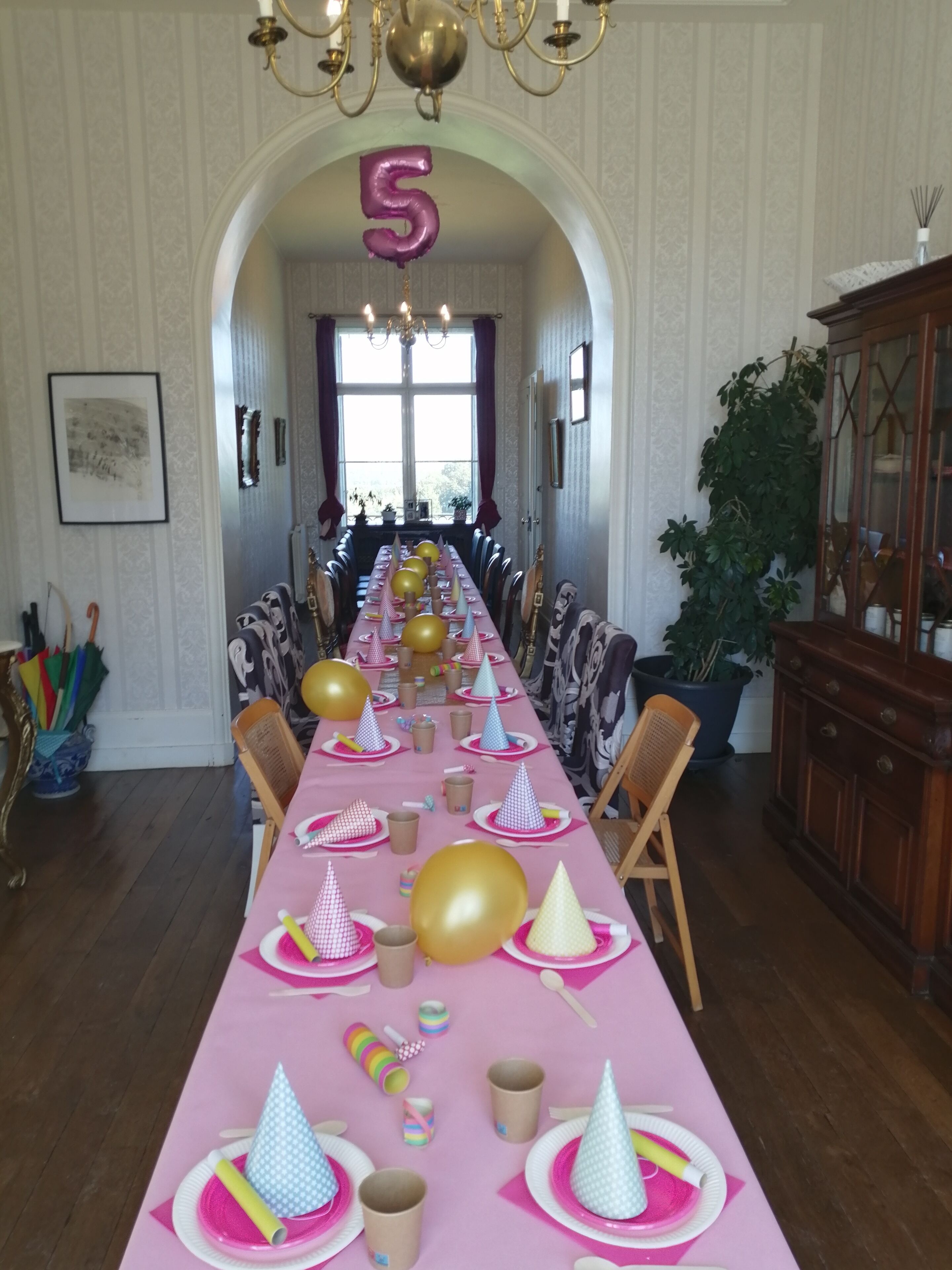 A long dining table set for a party with party hats, balloons, and a number '5' balloon.