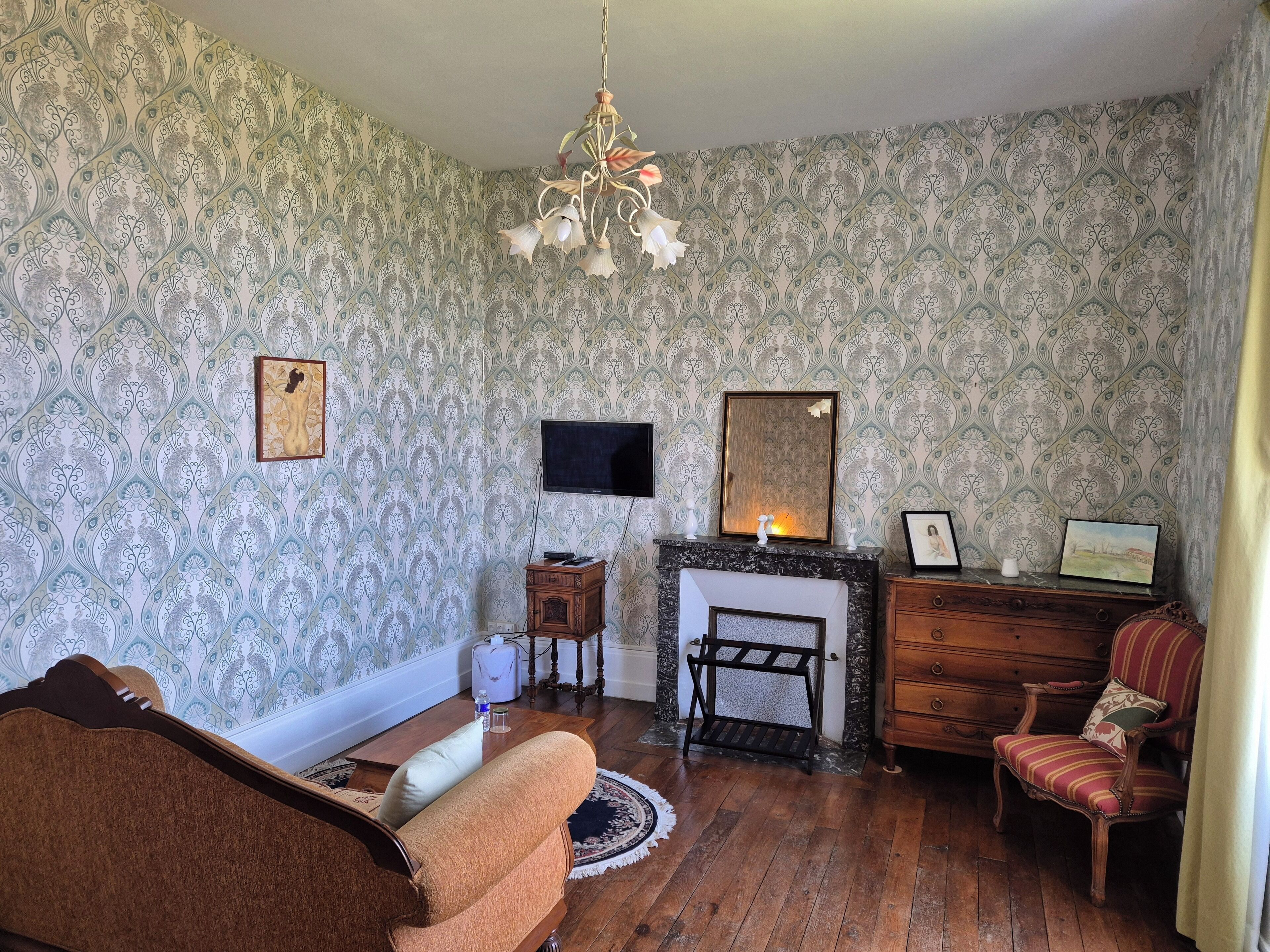 Un salon chaleureux avec un papier peint à motifs, une cheminée, une table basse en bois, un canapé, un fauteuil et un lustre.