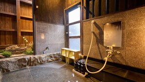 A modern bathroom with a stone wall, wooden paneling, a window, a shower area, and a small seating area. - Kigannoyado Aoshima Jizoan (Miyazaki)