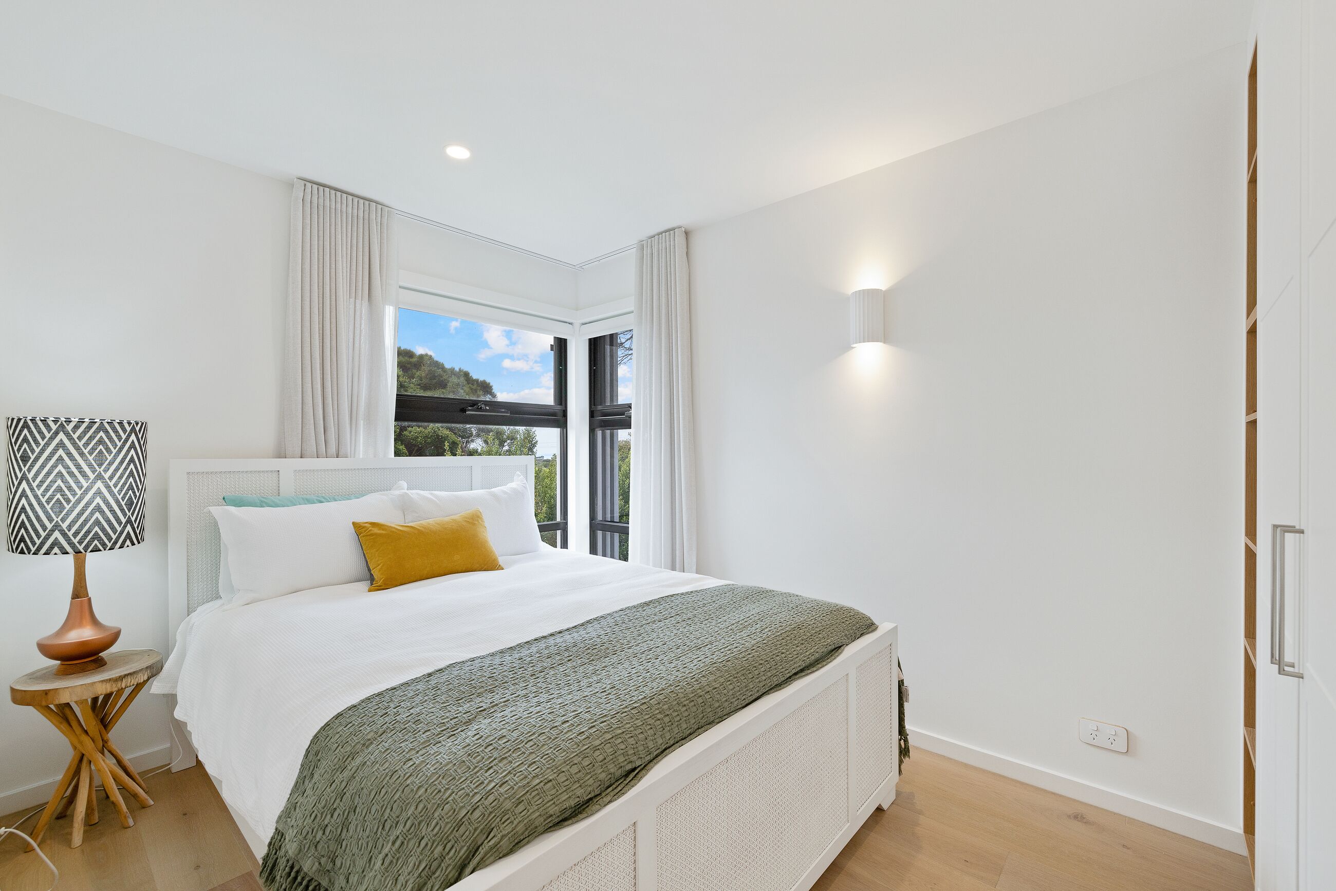 A modern bedroom with a large bed, a bedside lamp, and a view of the outdoors.