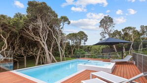 Une piscine dotée d’une terrasse en bois, de chaises longues et d’une structure en verre, le tout entouré d’arbres.