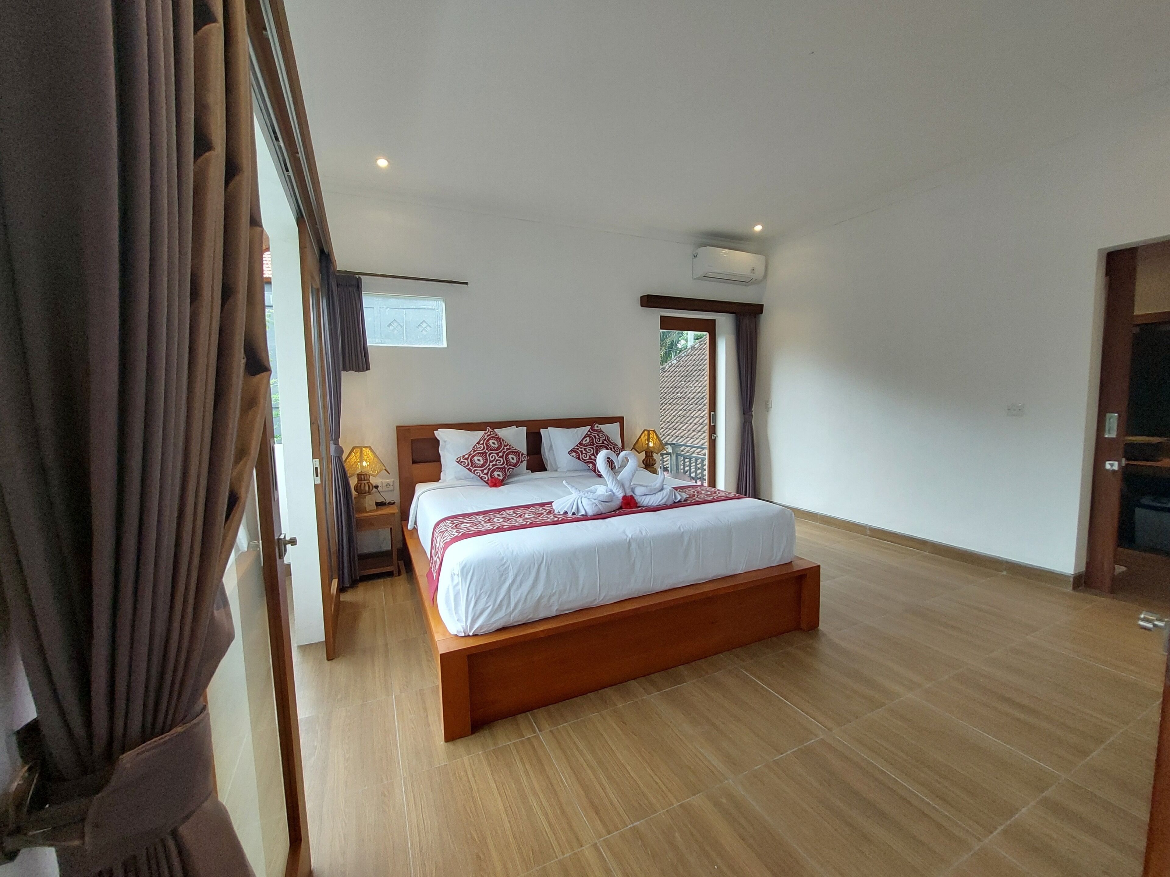 A hotel room with a wooden bed, a nightstand with a lamp, a ceiling fan, and a view of the outside.