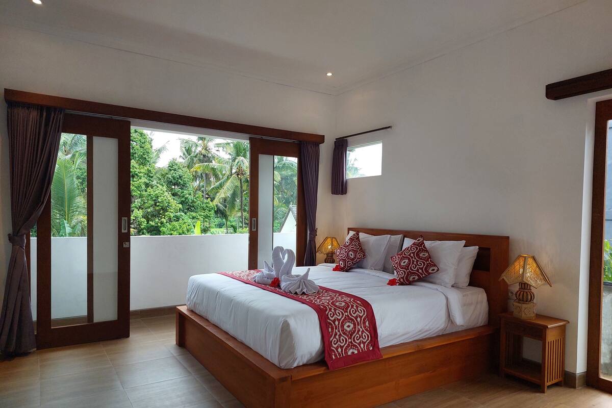 Ruime slaapkamer met een groot bed, houten hoofdbord en uitzicht op weelderige begroeiing door de schuifdeuren van glas.