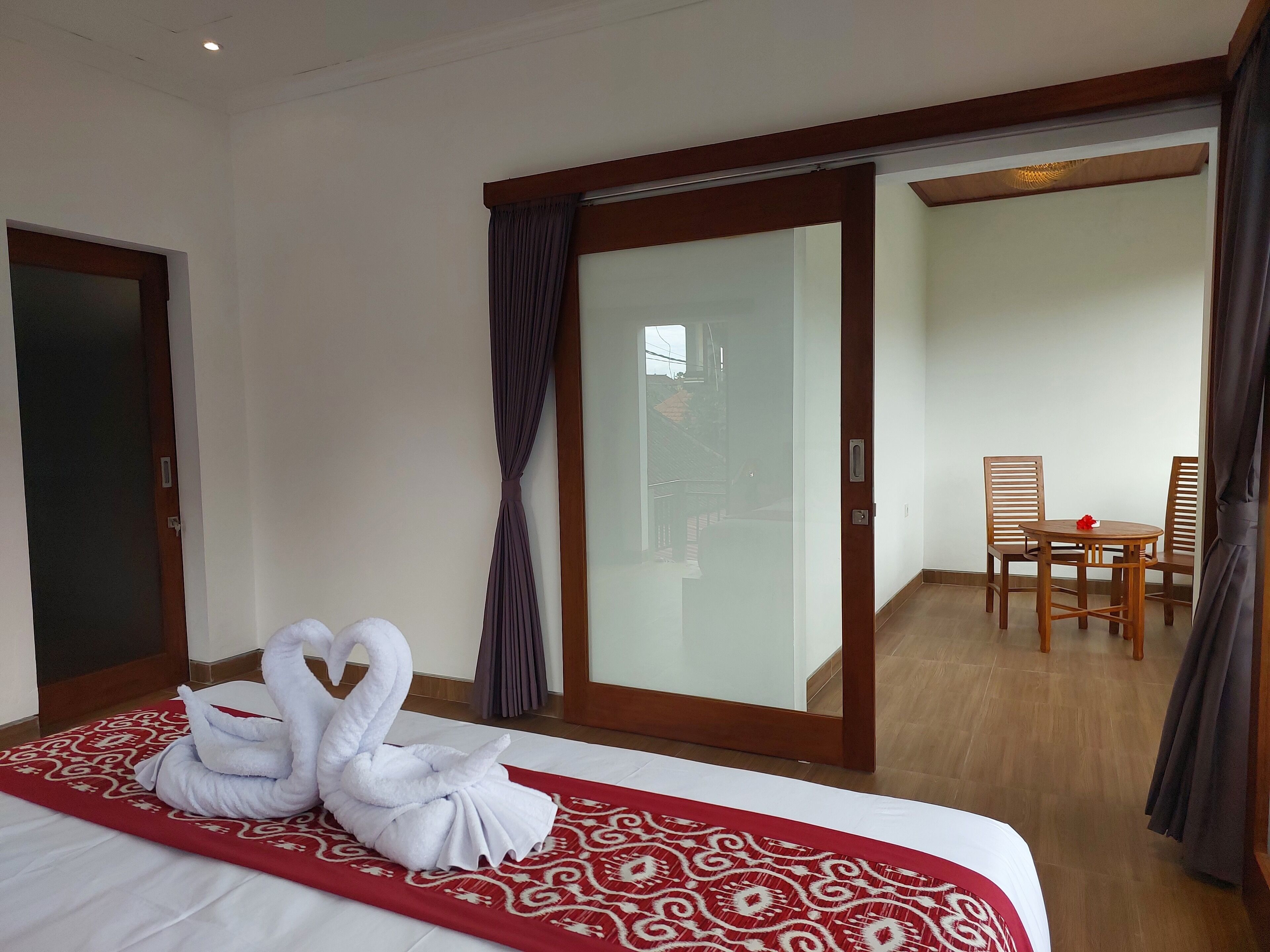 A hotel room with a bed, a red patterned bedspread, a swan towel arrangement, a sliding glass door, and a dining area with a table and chairs.
