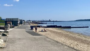 Een kustpad met een zandstrand, een heldere lucht en een industriegebied in de verte.