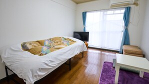 A small, well-lit room with a bed, a television, a window with curtains, and a table. - Aoshima Loco Beach (Miyazaki)