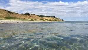 Vista clara de uma praia com águas rasas, areal arenoso e uma colina com vegetação rasteira ao fundo.