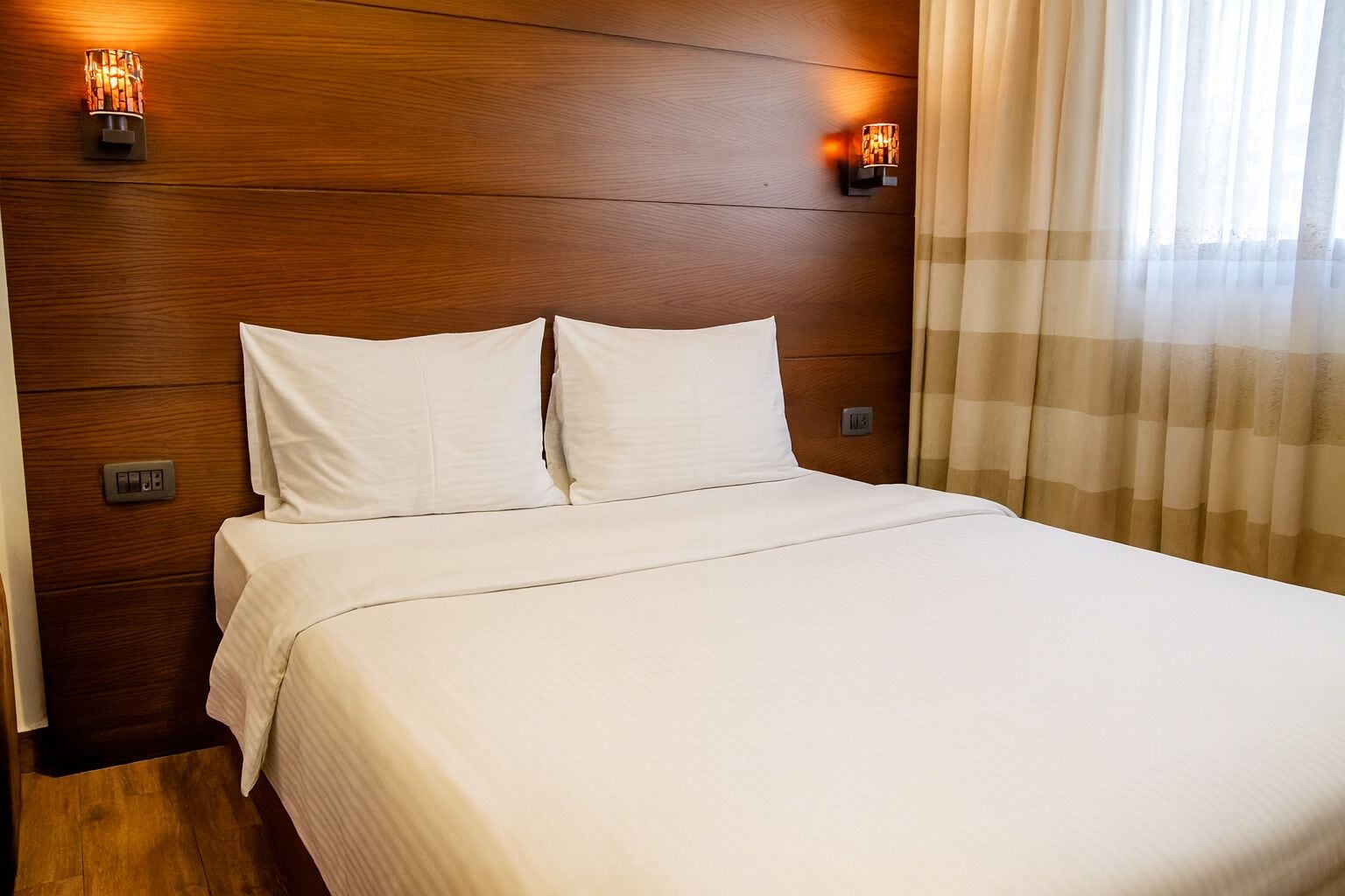 Una cama bien hecha con sábanas blancas en una habitación con paneles de madera y dos lámparas fijadas a la pared.