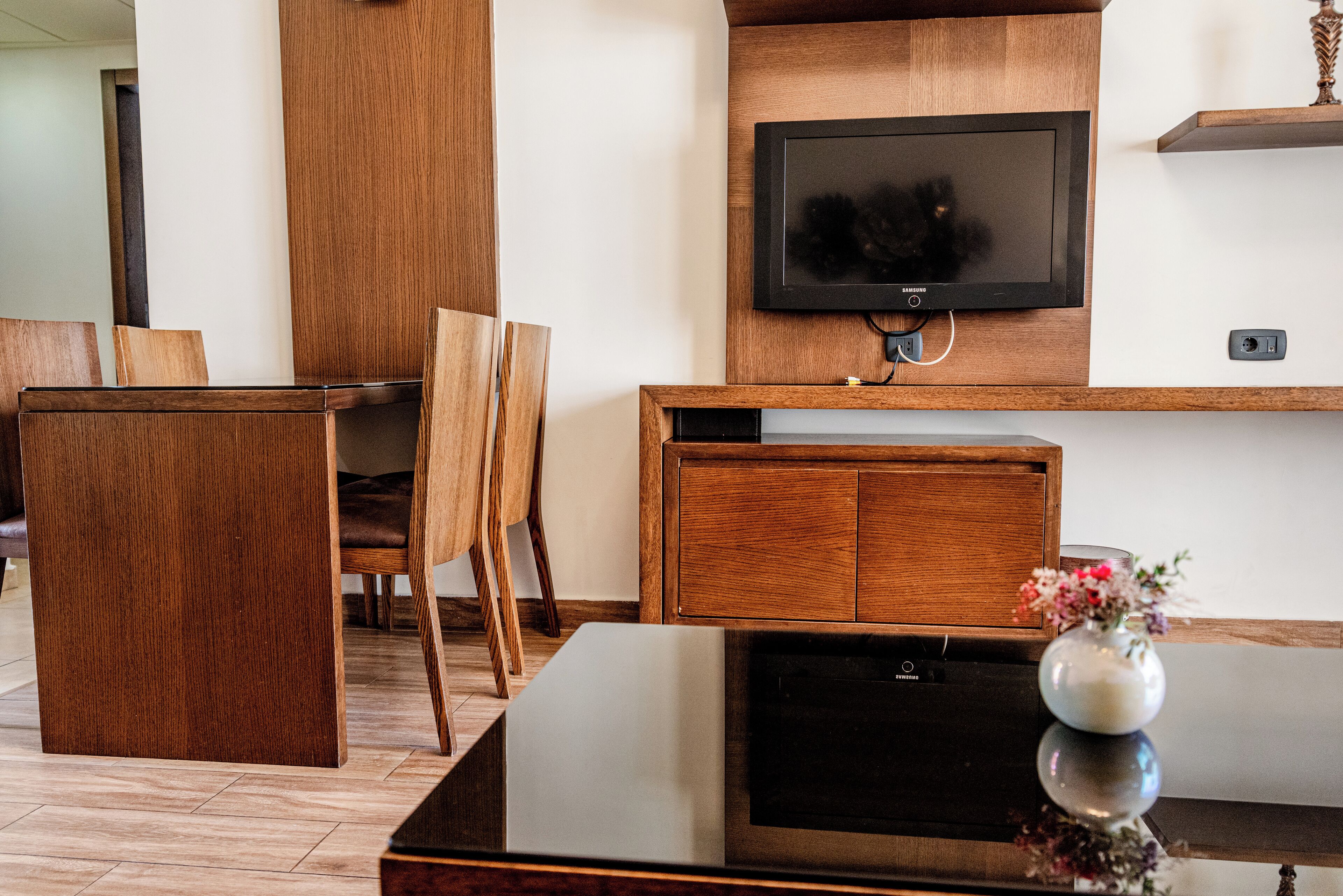 A modern living room with a flat-screen TV mounted on a wooden wall, a glass coffee table, and a wooden cabinet.