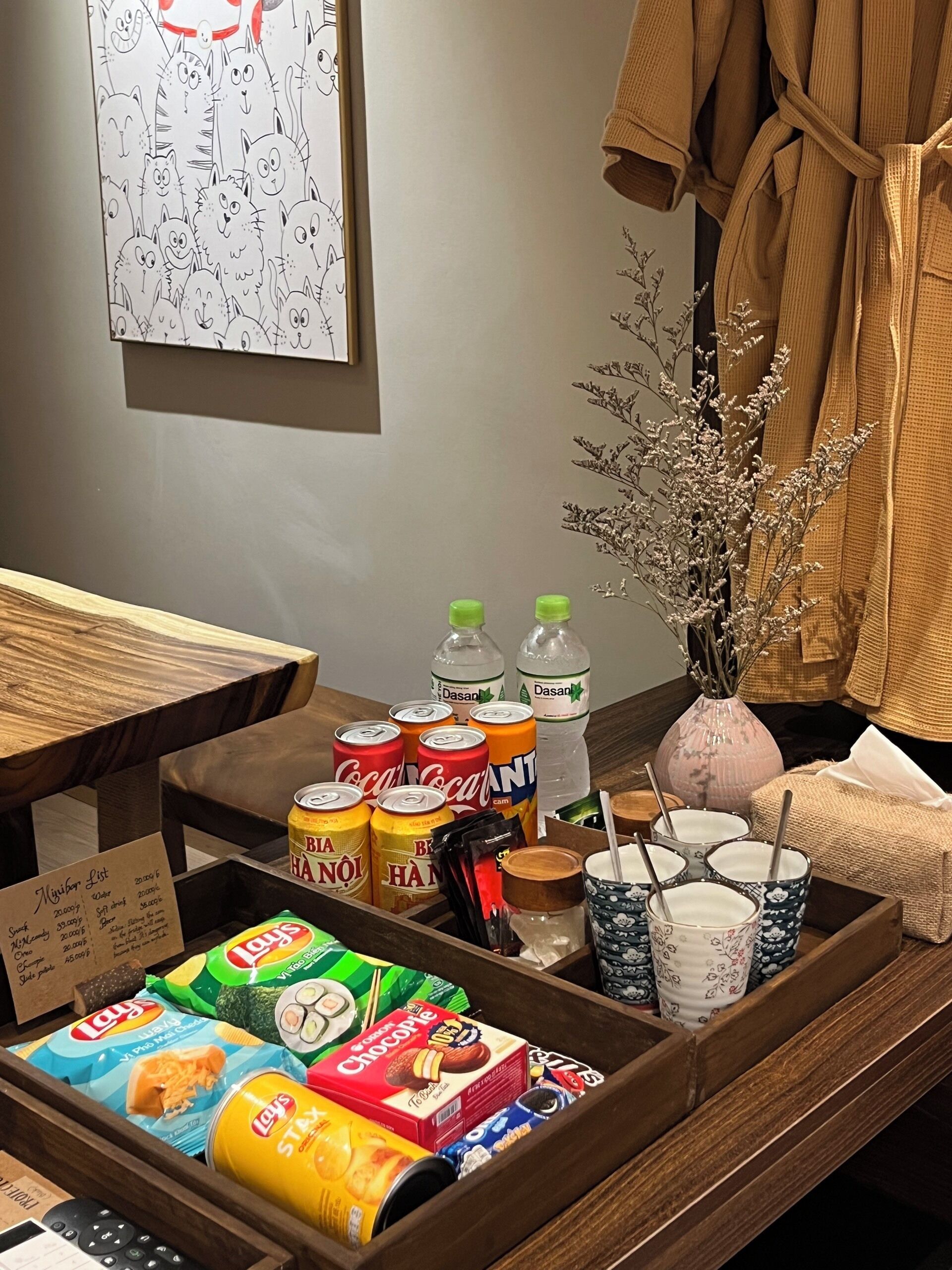A hotel room with a wooden side table holding snacks, drinks, and a vase with dried flowers.