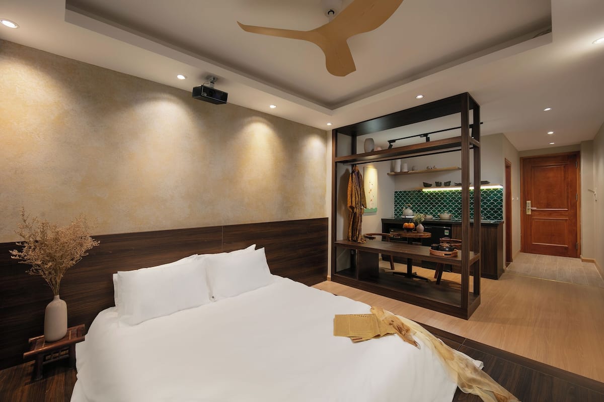 Ein modernes Hotelzimmer mit einem großen Bett, einem Holzkopfende, einem Deckenventilator, einem kleinen Tisch mit einer Vase getrockneter Blumen und Blick in den Küchenbereich.