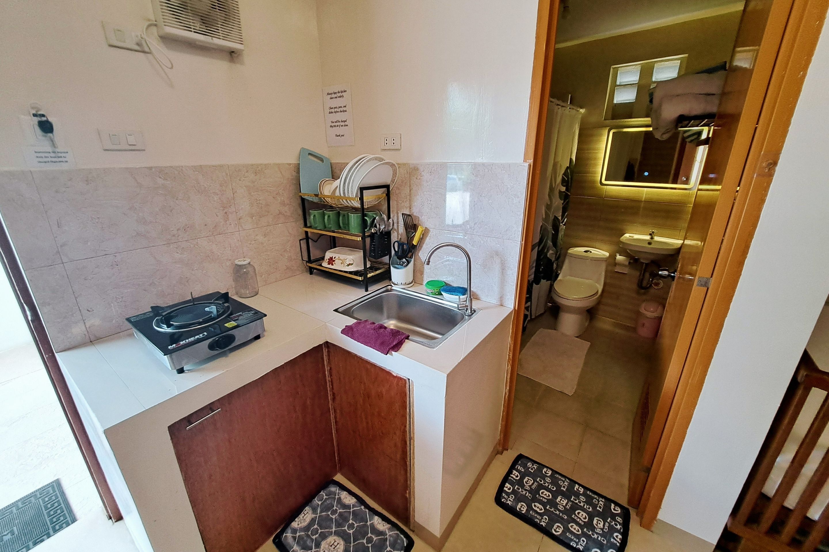 Une cuisine compacte équipée d’une cuisinière à gaz, d’un évier et d’un égouttoir. Une petite salle de bain avec des toilettes et un lavabo est visible à travers une porte ouverte.