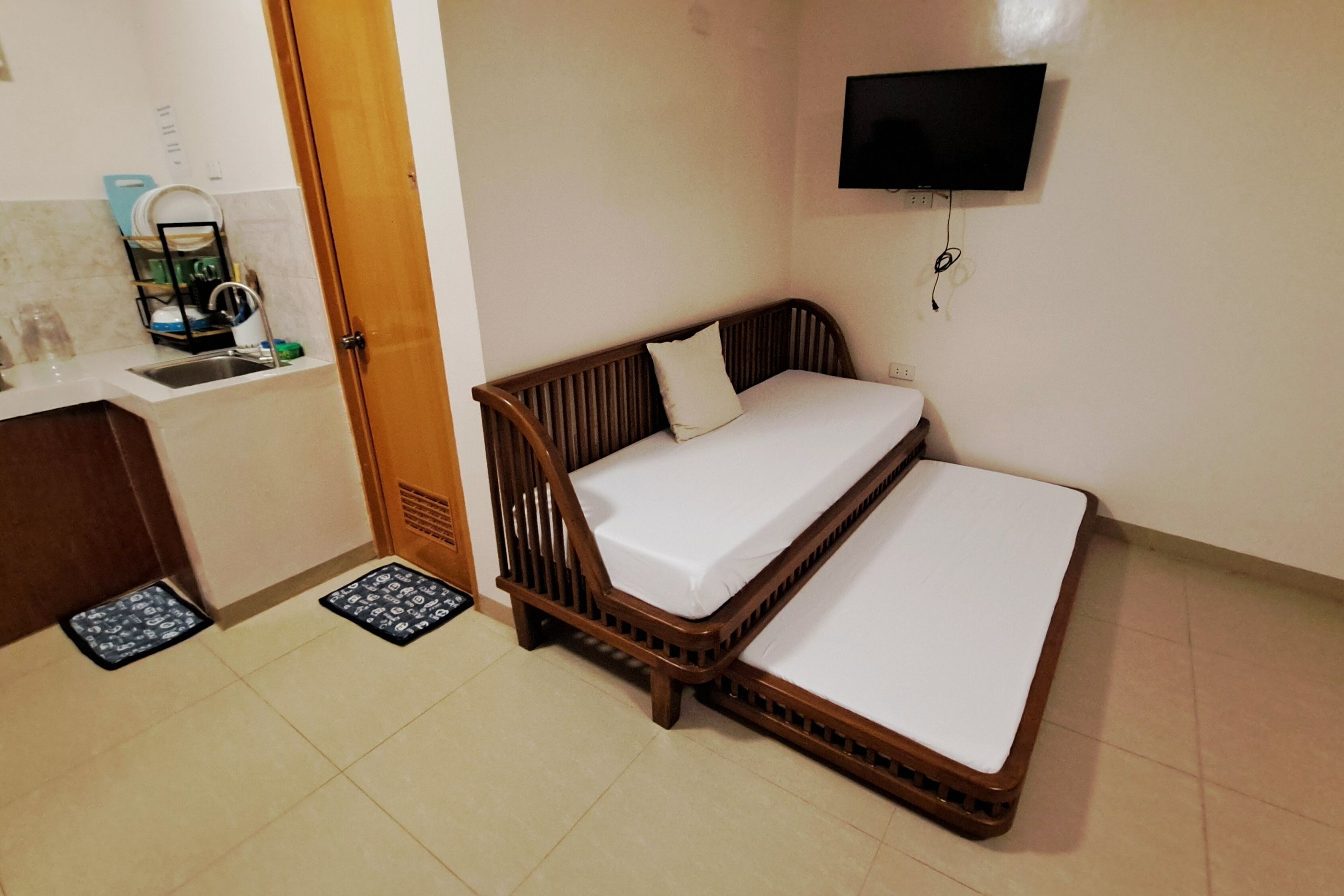 A small, compact room with a wooden bed, a television mounted on the wall, and a kitchen area with a sink and dish rack.