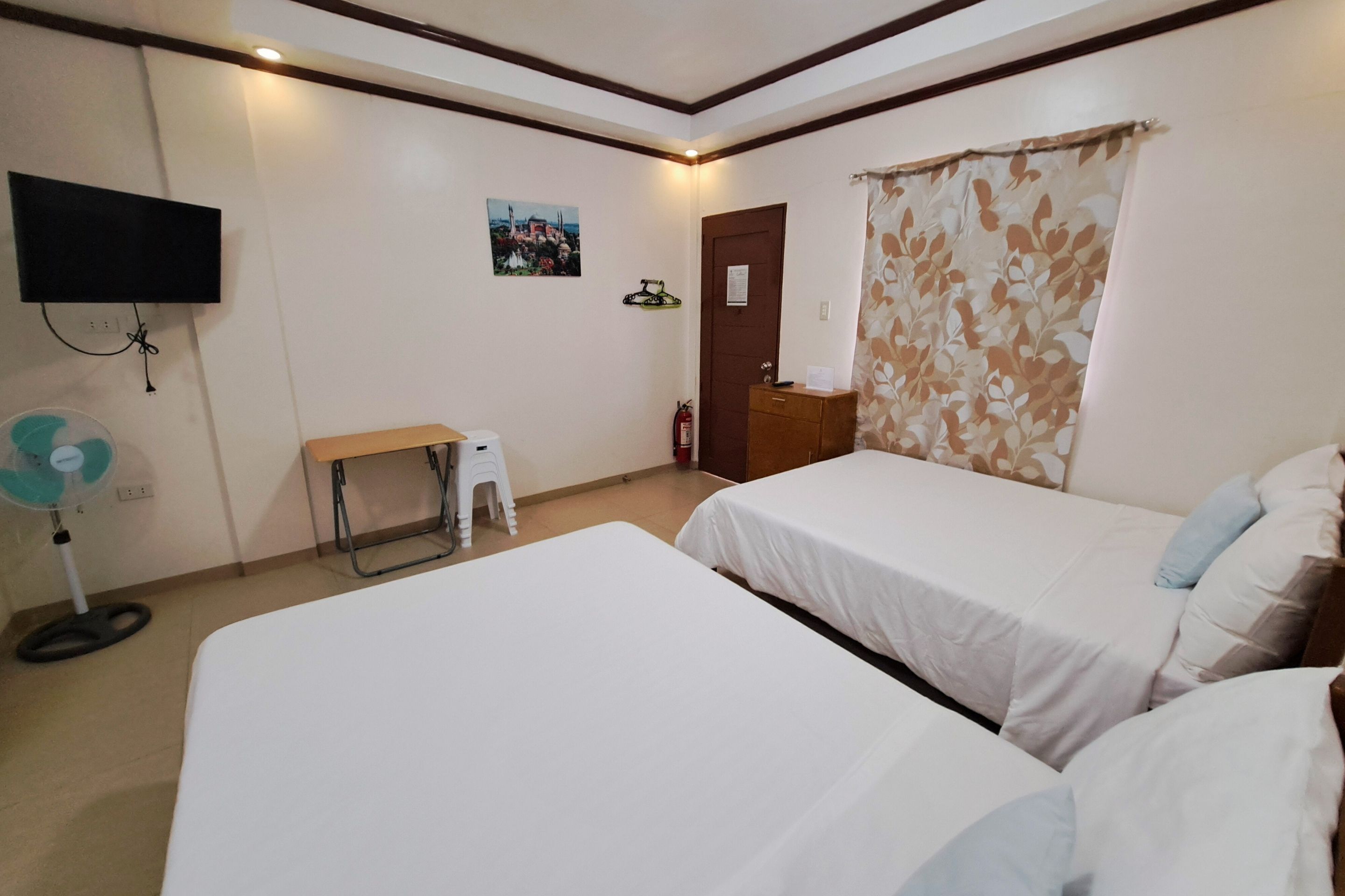 Habitación de hotel con dos camas, un televisor en la pared, una mesita, un ventilador y una puerta con cortina.