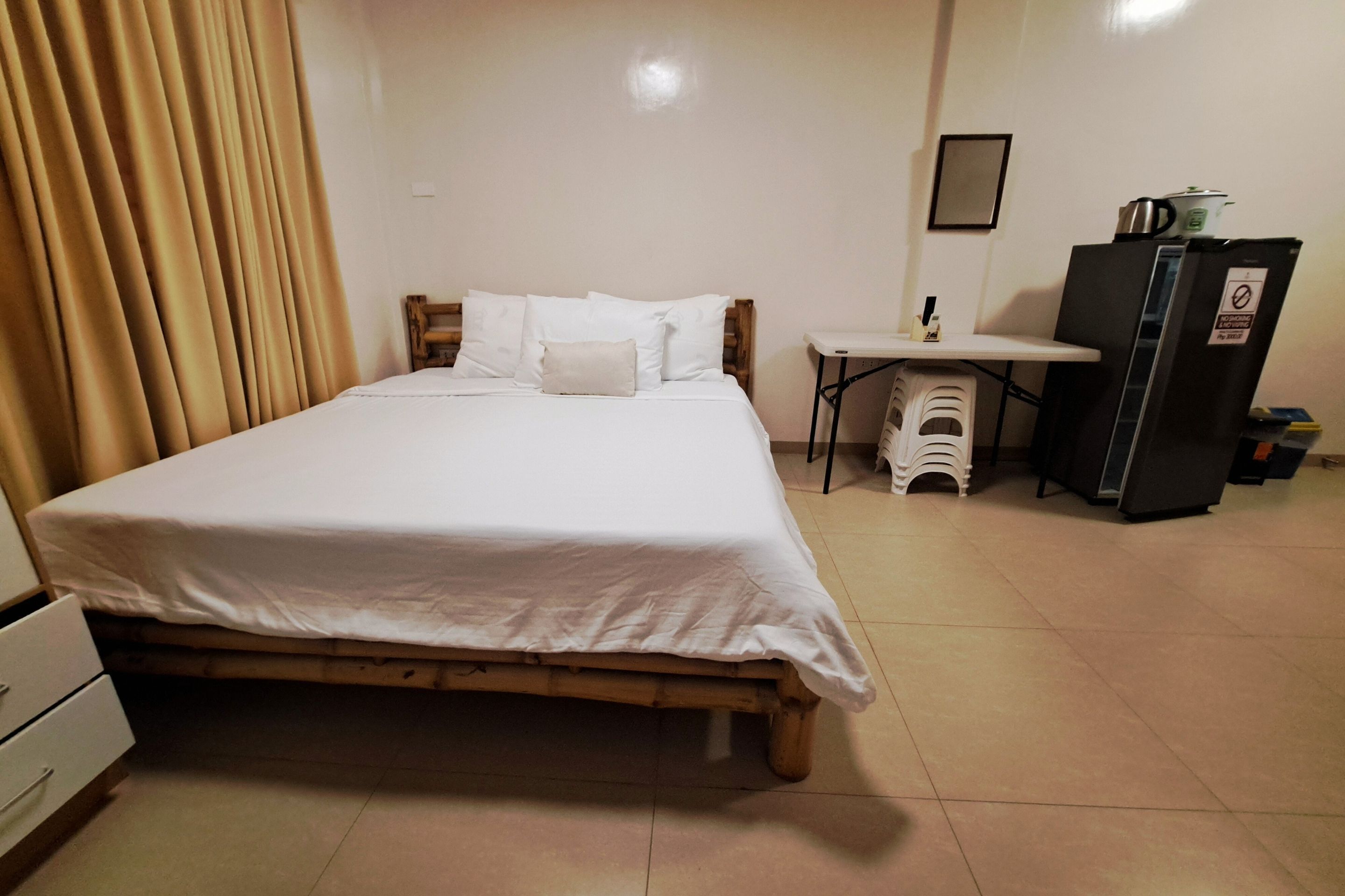 A neatly arranged bedroom with a wooden bed, white bedding, a small table, a refrigerator, and a white cabinet.
