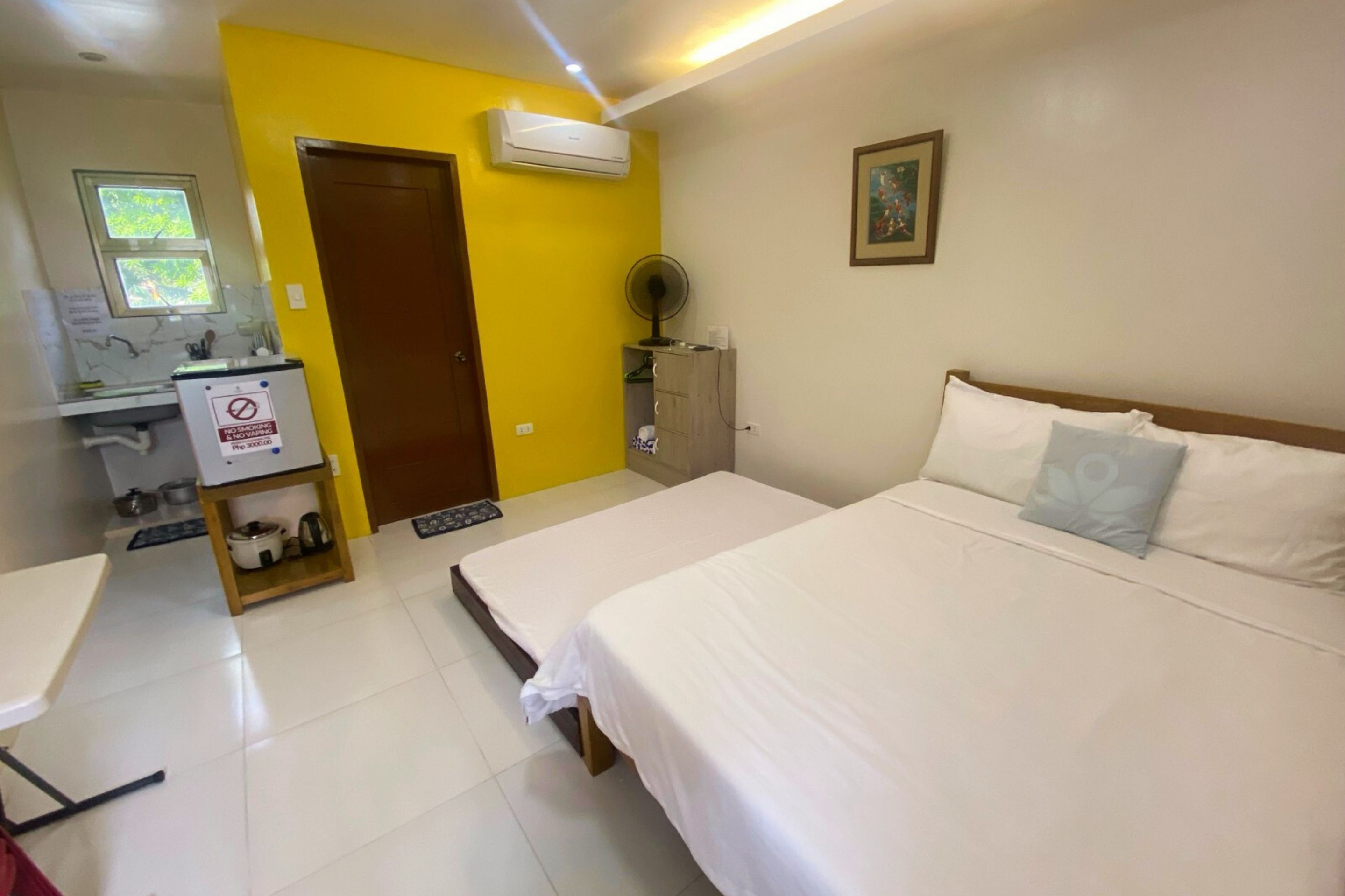 A hotel room with a bed, a desk, a chair, a small kitchenette, and a door.