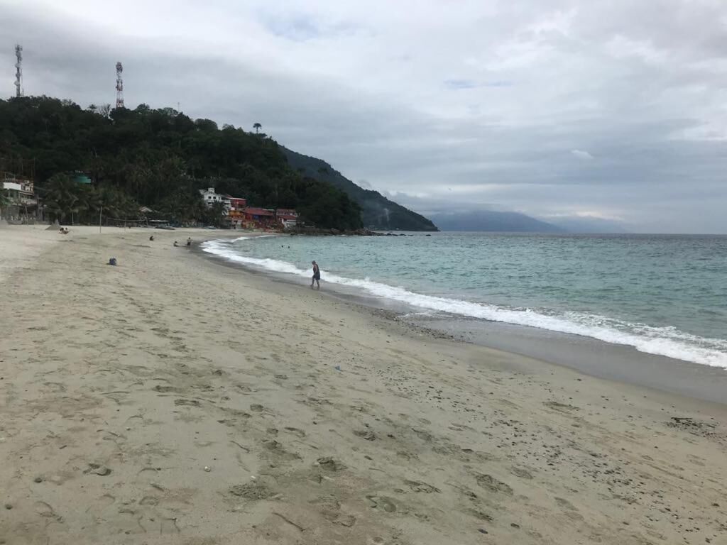 A beach with a sandy shore, a few people walking, and buildings on a hillside in the background.
